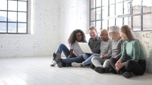 Cheerful multiracial creative coworkers in casual clothes resting on floor while using netbook and working on project together in loft styled room