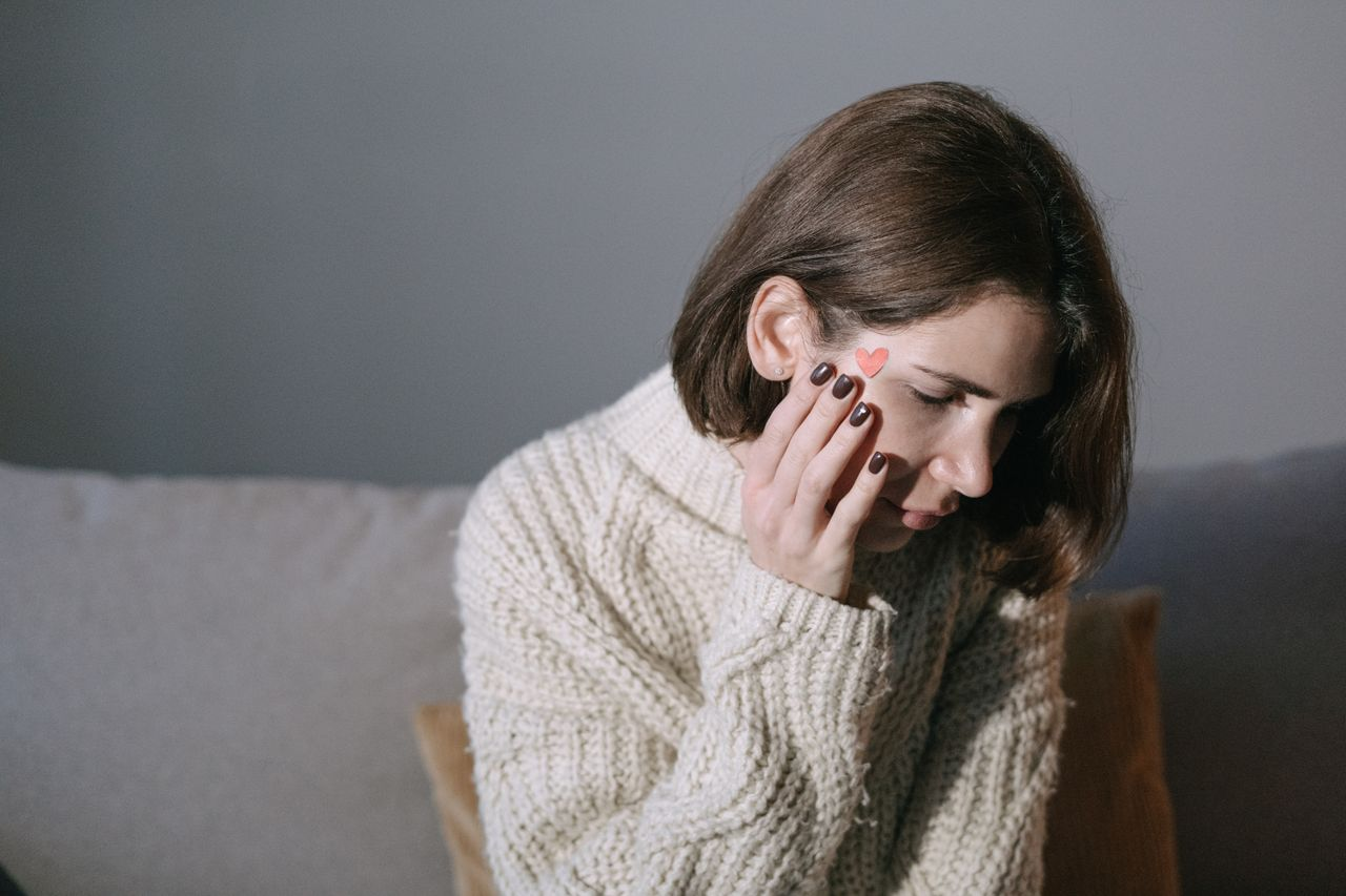 Mastering the First Impression: Your intriguing post title goes here A woman wearing a sweater rests her hand on her face, displaying a heart sticker, in a softly lit room.