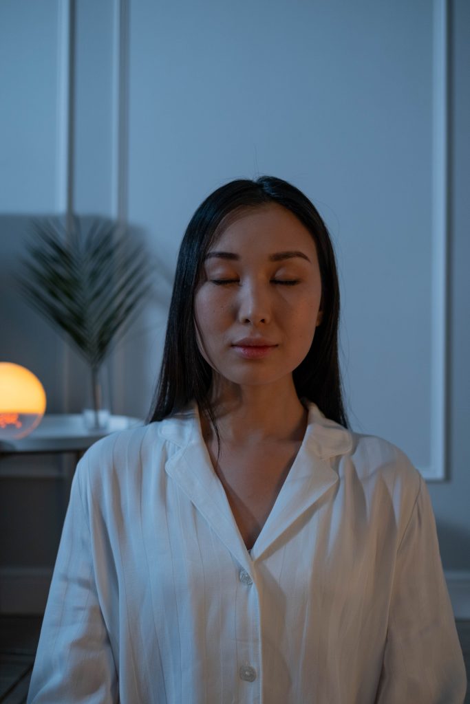 Close-up portrait of a woman with eyes closed, wearing white pajamas, in a calm indoor setting.